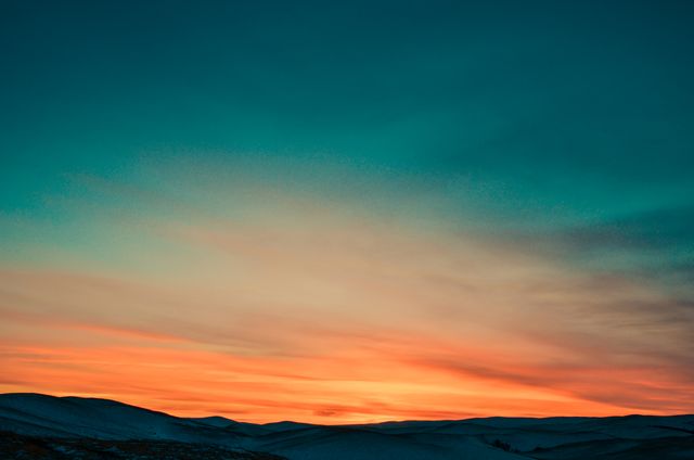 An image capturing a vibrant sunset over gently rolling hills in a desert landscape. The sky displays a gradient of warm colors ranging from orange to teal, offering a tranquil and calming scene. Suitable for website backgrounds, travel magazines, nature portfolios, relaxation and meditation apps, or any project requiring a peaceful and scenic backdrop.