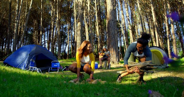 Group of friends enjoying a camping trip in the forest. Two friends are setting up a campfire while another sits in the background playing the guitar. Tents are set up around them, indicating it's a campsite. This image can be used for outdoor adventure themes, camping advertisements, travel blogs, and friendship or bonding concepts.