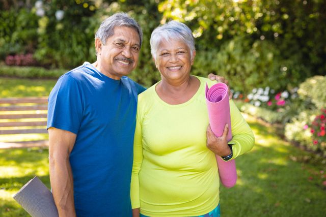 Portrait of smiling biracial senior couple with exercise mats standing against plants in park. unaltered, love, togetherness, retirement, fitness and active lifestyle concept.