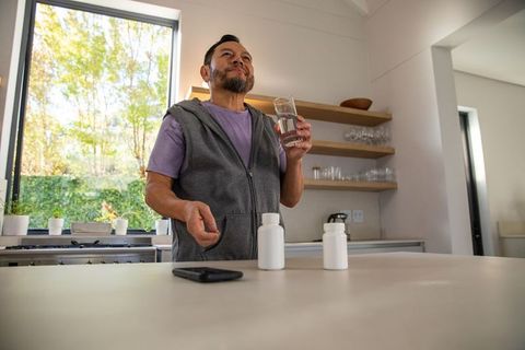 Senior asian man taking daily medication with glass of water in bright modern kitchen