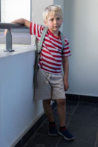 Thoughtful schoolboy leaning on balcony railing with backpack and red striped shirt