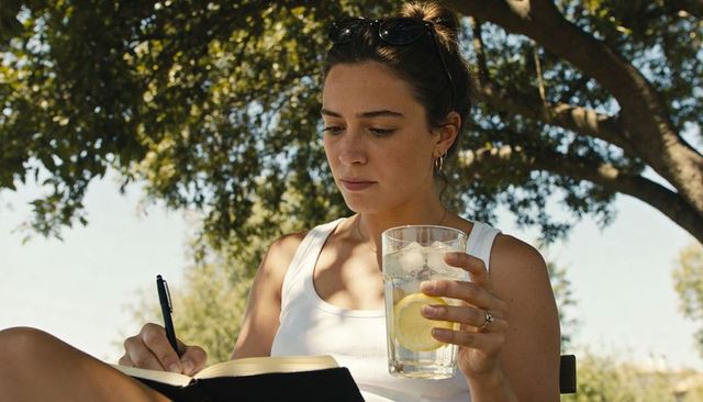 Young woman writing in journal on park bench holding lemon water under oak tree