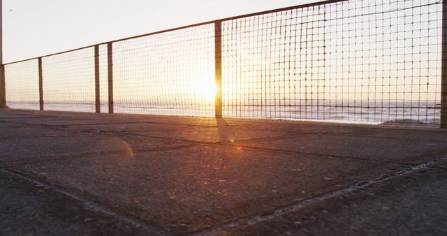 Sunset casting golden rays through wire fence along oceanfront promenade, creating tranquil scene with shadows and textures on pavement. Ideal for content related to travel, relaxation, coastal living, outdoor beauty, or atmospheric backdrops for presentations or websites.