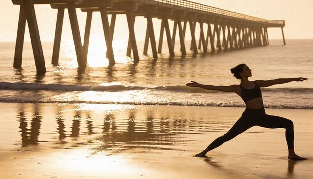 Woman practicing warrior ii yoga on sunlit wet sand by pier with golden reflection at sunrise