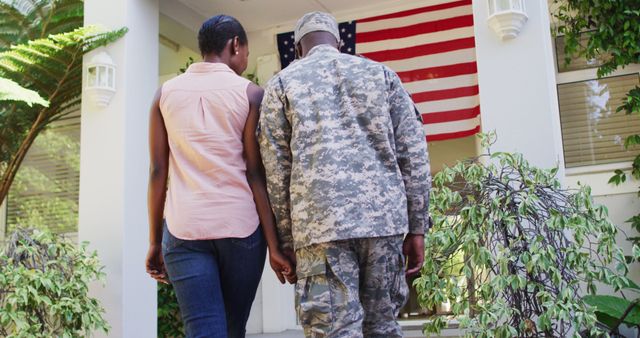 Happy african american male soldier and wife holding hands walking to house with american flag. soldier returning home to family.