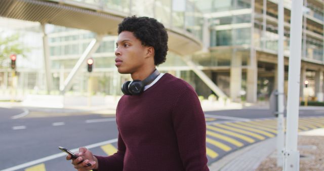 Young man wearing headphones walking across a crosswalk in a modern urban area while checking his smartphone. Ideal for campaigns related to modern technology, urban lifestyle, youth culture, or transportation safety.