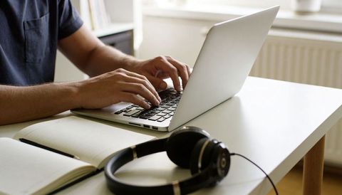 Man typing on laptop at minimalist home desk with notebook and headphones for remote work
