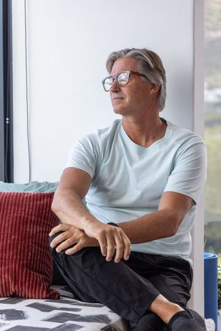 Middle-aged man sitting on window seat gazing out window wearing tortoiseshell glasses