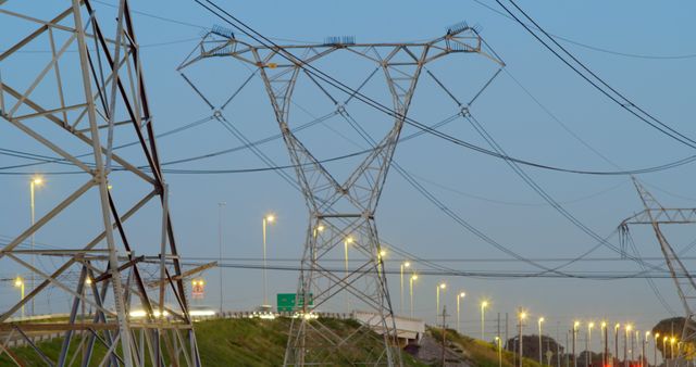 Power lines stretching across the image at dusk along a highway, with lights illuminating the area and vehicles passing by. Useful for presentations about energy transmission, infrastructure projects, renewable energy concepts, and illustrating industrial landscapes.