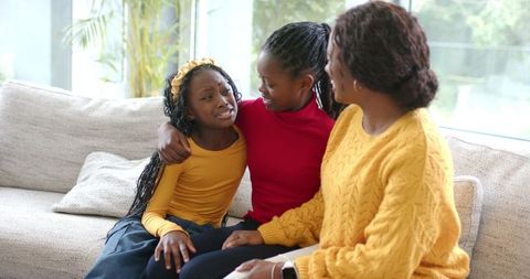 African American Mom Comforting Upset Daughter on Sunlit Sofa with Supportive Sister