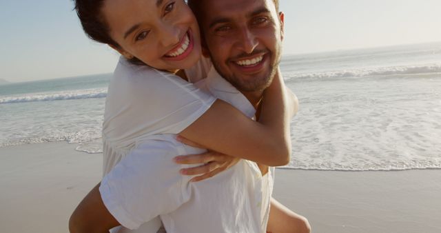 A joyful couple enjoying a piggyback ride on a sandy beach at sunset. Perfect for promoting travel destinations, romantic getaways, summer vacations, and relationship goals. Captures a sense of happiness, love, and carefree moments with the ocean waves in the background.