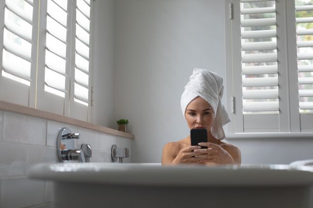 Woman sitting in a bathtub using a mobile phone with a towel wrapped around her hair. Ideal for concepts related to relaxation, self-care, modern lifestyle, and technology use in everyday life. Can be used in advertisements for bath products, mobile apps, or home decor.