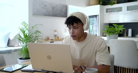 Young man working remotely at bright modern kitchen table wearing headphones and beanie