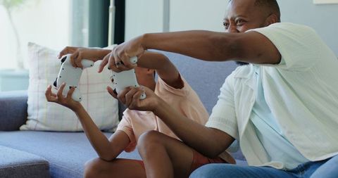 Father and son competing with wireless game controllers on sofa, playful family bonding