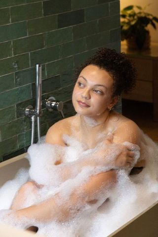 African american woman soaking in bubble bath freestanding tub dark green tile backdrop