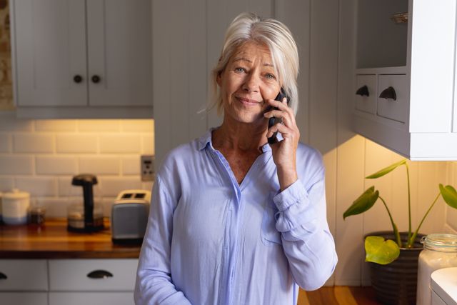 Smiling mature caucasian woman standing in kitchen making phone call on smartphone, copy space. Domestic life, living alone, communication and senior lifestyle concept.