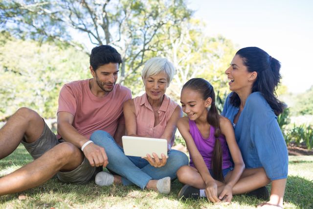 Family members of different generations are sitting on grass in a park, enjoying time together while looking at a digital tablet. This image can be used for promoting family bonding, outdoor activities, technology in daily life, and leisure time. Ideal for advertisements, blogs, and articles related to family, technology, and outdoor recreation.