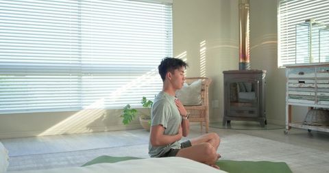 Asian man practicing mindful breathing on green mat in sunlit minimalist living room