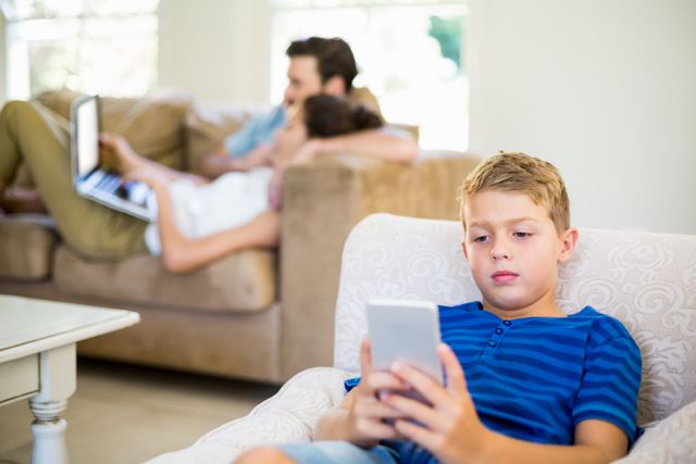 Boy sitting on sofa using mobile phone, with parents in background using laptops. Ideal for illustrating modern family life, digital device usage, and home relaxation. Suitable for articles on technology impact on children, family dynamics, and home environments.