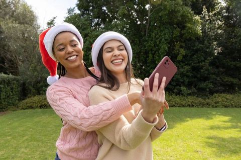 Diverse friends wearing santa hats hugging and taking selfie in sunlit park holiday joy