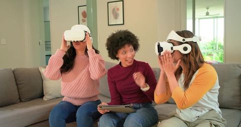 Diverse women using VR headsets on sofa sharing tablet in bright modern living room