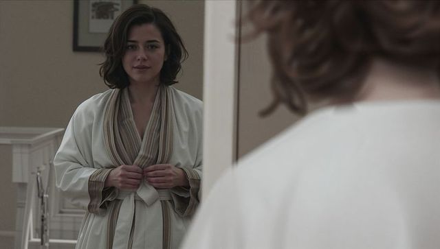 Tying cream robe with striped trim, woman facing mirror in minimalist beige hallway