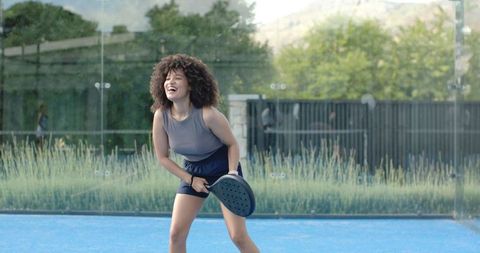 Joyful african american woman playing pickleball on blue court laughing with paddle