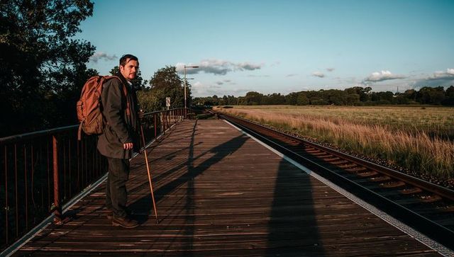 Solo traveler standing on rural platform with backpack and walking stick at golden hour