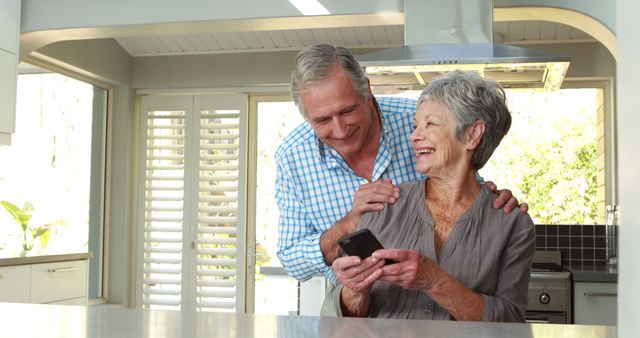 Senior couple smiling and enjoying time together in modern kitchen while using smartphone. Can be used for topics related to senior living, modern technology, communication, family bonding, and promoting positive and active aging among retirees.