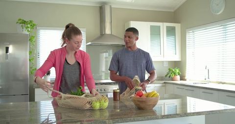 Couple unpacking reusable mesh grocery bags on modern kitchen island with fresh produce