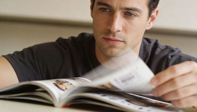 Man flipping glossy magazine at lounge table, contemplative reading moment with soft light