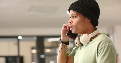 African american man talking on smartphone with headphones and beanie in modern coworking office