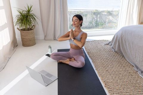 Practicing morning meditation on yoga mat in sunlit bedroom with laptop
