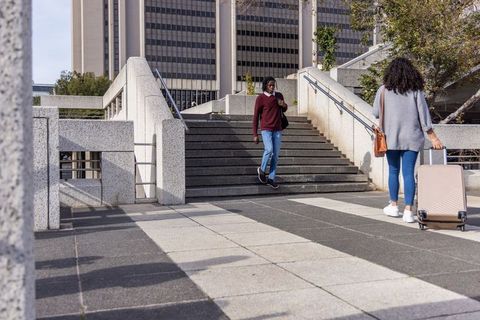 African american man descending stairs while woman pulling suitcase across urban plaza