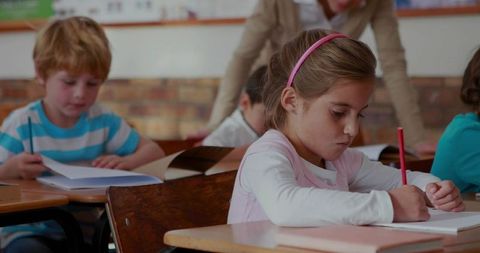 Young girl writing with pink headband concentrating on red pencil at wooden school desk