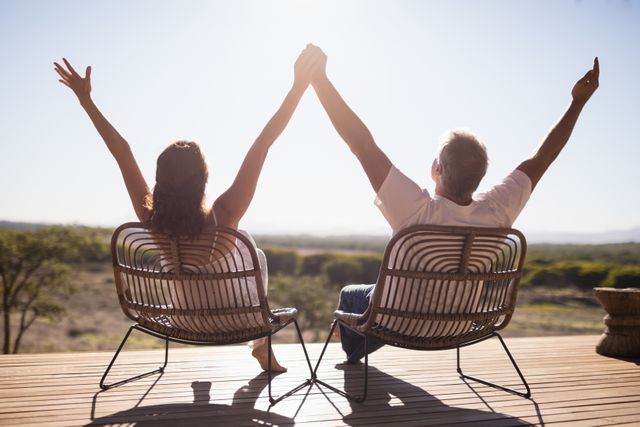 Senior couple sitting on chairs outdoors with arms raised, enjoying leisure time at a resort. Ideal for use in advertisements for retirement communities, travel agencies, wellness retreats, or lifestyle blogs focusing on senior living and relaxation.