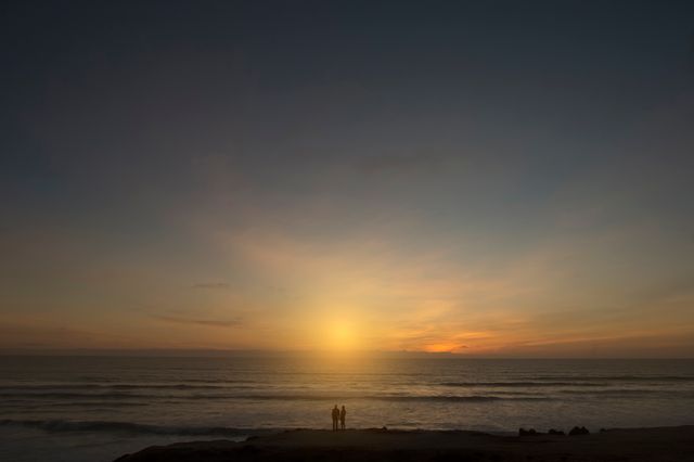 Couple stands on beach watching sun set over calm ocean. Sky lit with warm colors, creating peaceful and romantic atmosphere. Ideal for themes of romance, leisure, travel inspiration, peaceful moments, nature appreciation, and couple bonding.