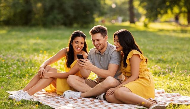 Three friends enjoying golden-hour picnic, sharing smartphone laughs on checkered blanket