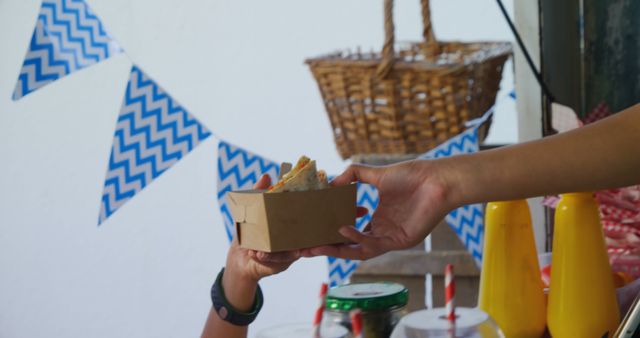 Person in outdoor fair booth handing cardboard tray with sandwich to customer. Ideal for marketing street food events, food festivals, farmers' markets, or small business promotions in casual dining settings.