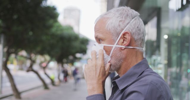 Senior man adjusting a protective face mask while standing outside in an urban area, emphasizing health and safety measures in public spaces. Suitable for content related to pandemic safety protocols, urban life, senior health care, and prevention of illness.