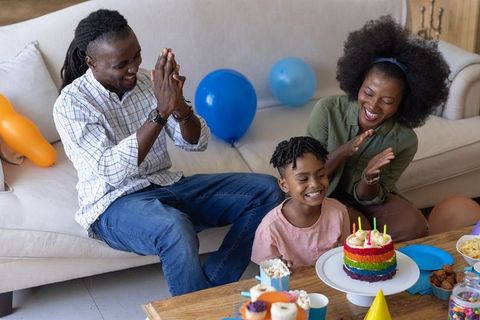 African american family celebrating child birthday at home with rainbow cake and balloons