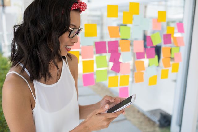 Female executive standing in modern office, using mobile phone while smiling. Background features colorful sticky notes on glass wall, indicating brainstorming or planning session. Ideal for business, technology, communication, and workplace productivity themes.
