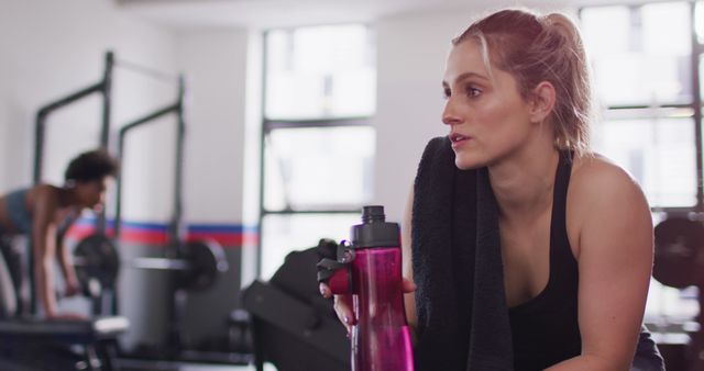 Woman is taking a rest while holding a water bottle during a gym workout. She appears to be in a modern gym setting, reflecting an active and healthy lifestyle. This image can be used in promoting fitness centers, workout programs, health and wellness articles, or any content related to staying fit and healthy.