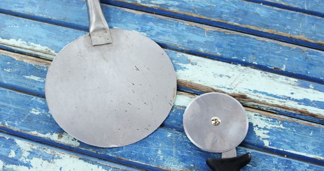 Metal kitchen utensils, including a large flat spatula and pizza cutter, placed on a weathered blue wooden plank table. Ideal for illustrating rustic kitchen settings, utensil advertising, culinary content, and kitchenware stores.