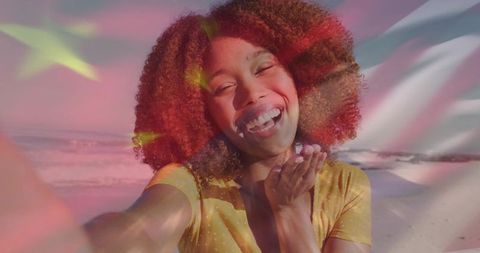 Sunlit beach selfie laughing woman with curly hair blowing kiss, flag overlay sunny glow