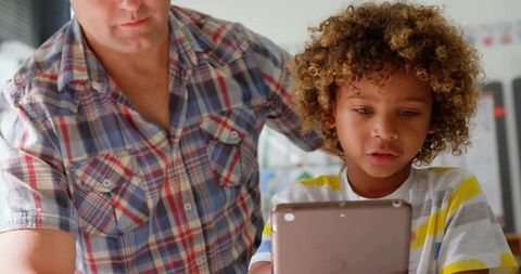 Mature father teaching curly-haired son with tablet at home