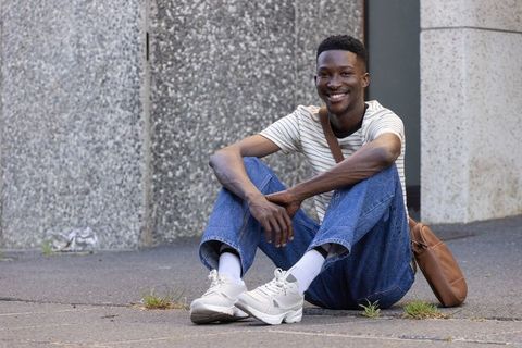 Young african american man sitting on city sidewalk smiling while holding leather satchel