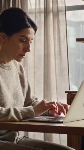 Vertical video of focused woman typing on silver laptop by sheer curtains for remote work