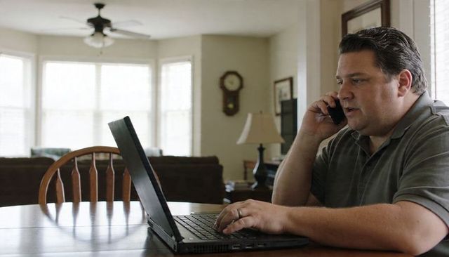 Man working from home on laptop while talking on phone at dining table with bay window natural light