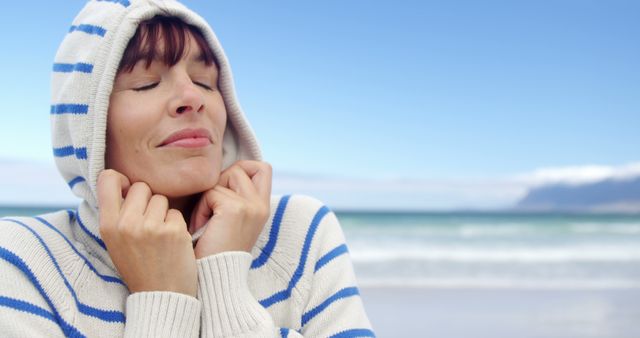 Woman stands on a beach, dressed in a striped hoodie, eyes closed, enjoying the calm seaside. Perfect for illustrating relaxation, nature enjoyment, coastal tourism, winter beach apparel, and personal tranquility.